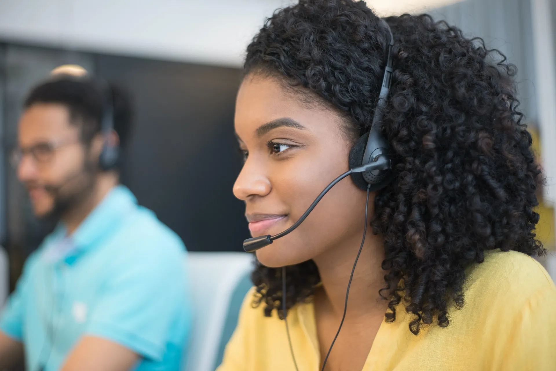 Customer service agent wearing a headset, with a colleague at a workstation in the background